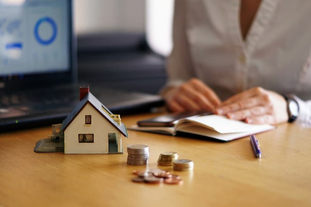Escritorio con figura de casa, monedas y mujer de fondo